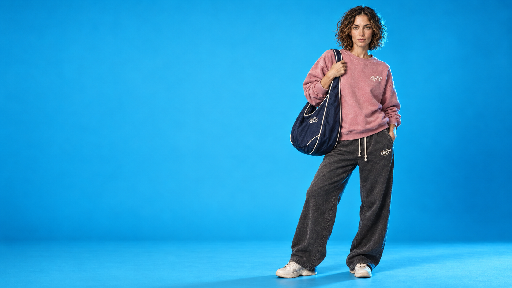 Lost Wight model wearing the faded rose sweatshirt and charcoal joggers with the navy Seafront bag against cyan blue