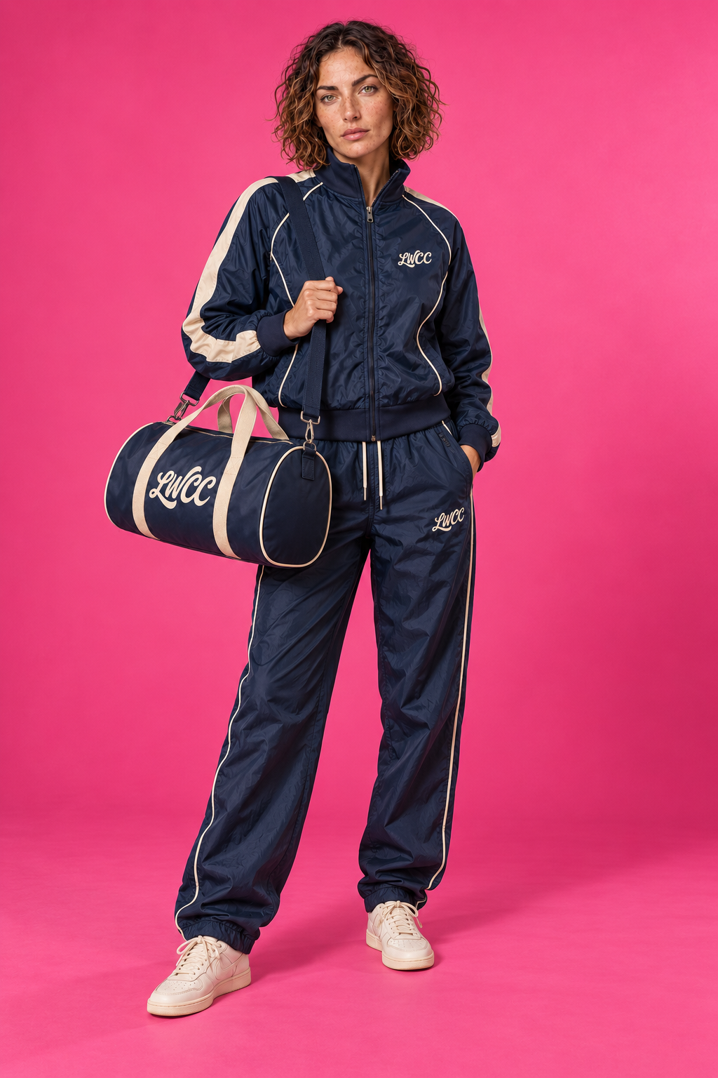 The same Lost Wight model wearing the navy and cream Court Piping shell jacket and matching trousers with the bowling bag over her shoulder, against neon pink