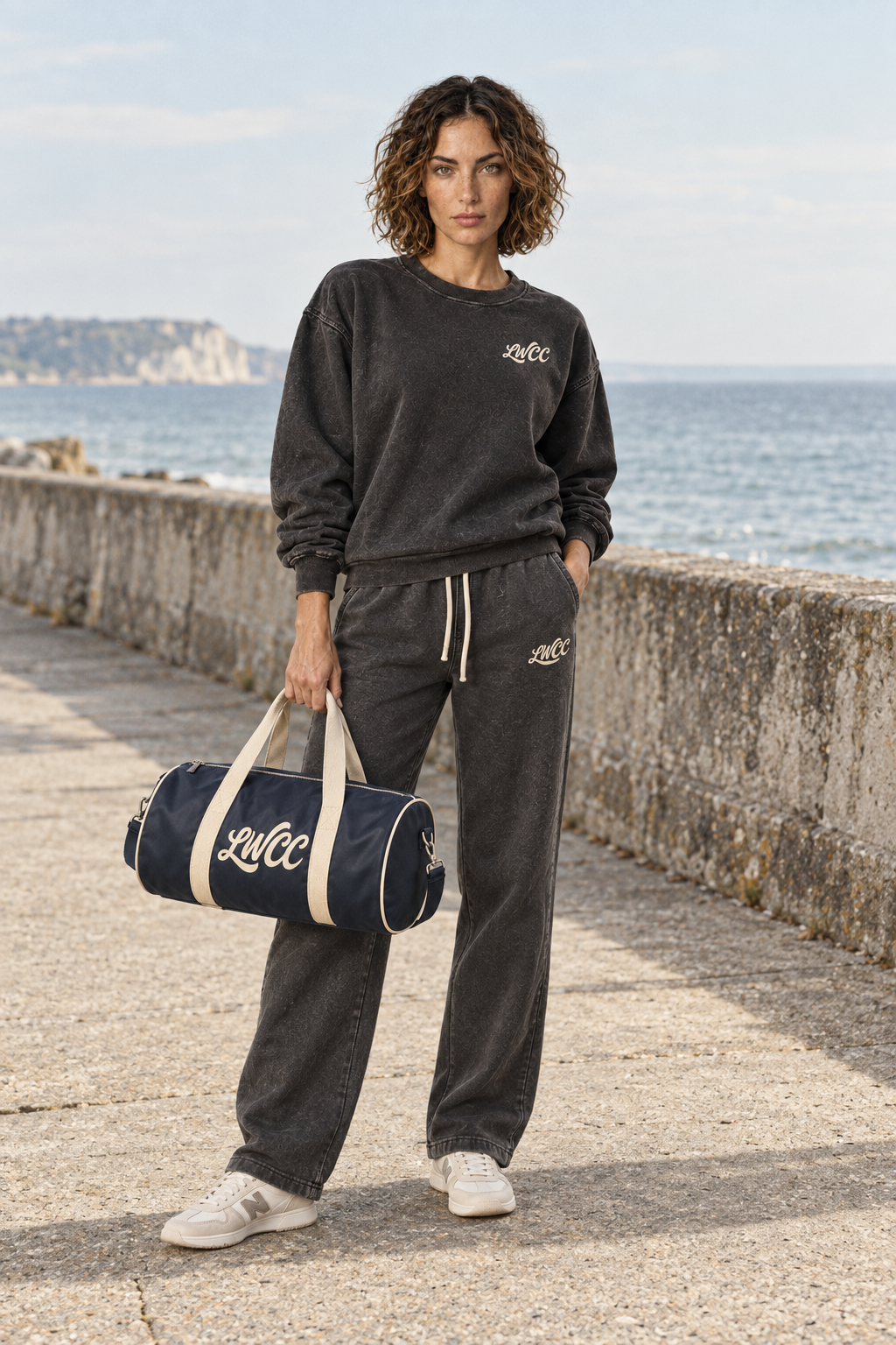 The Lost Wight model in washed charcoal sweatshirt and straight-leg joggers, carrying the navy and cream bowling bag on a seaside promenade